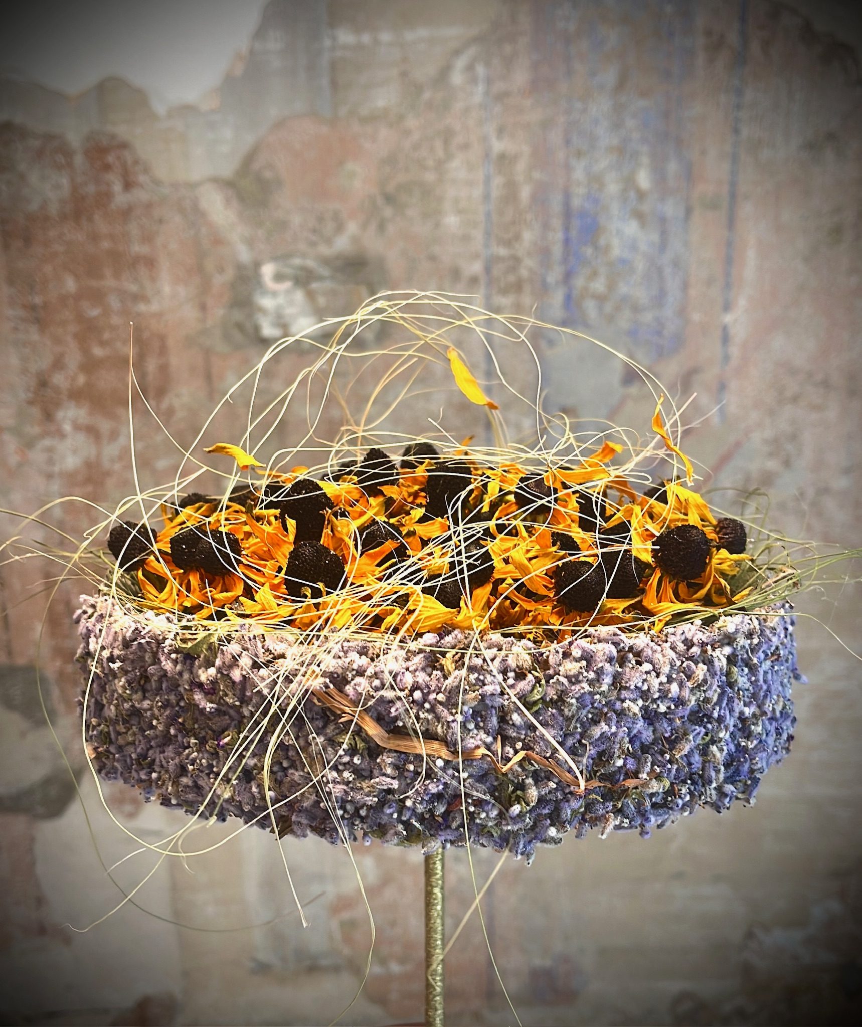 Scultura floreale di lavanda e calendula avvolta da fili dorati, davanti a una parete affrescata